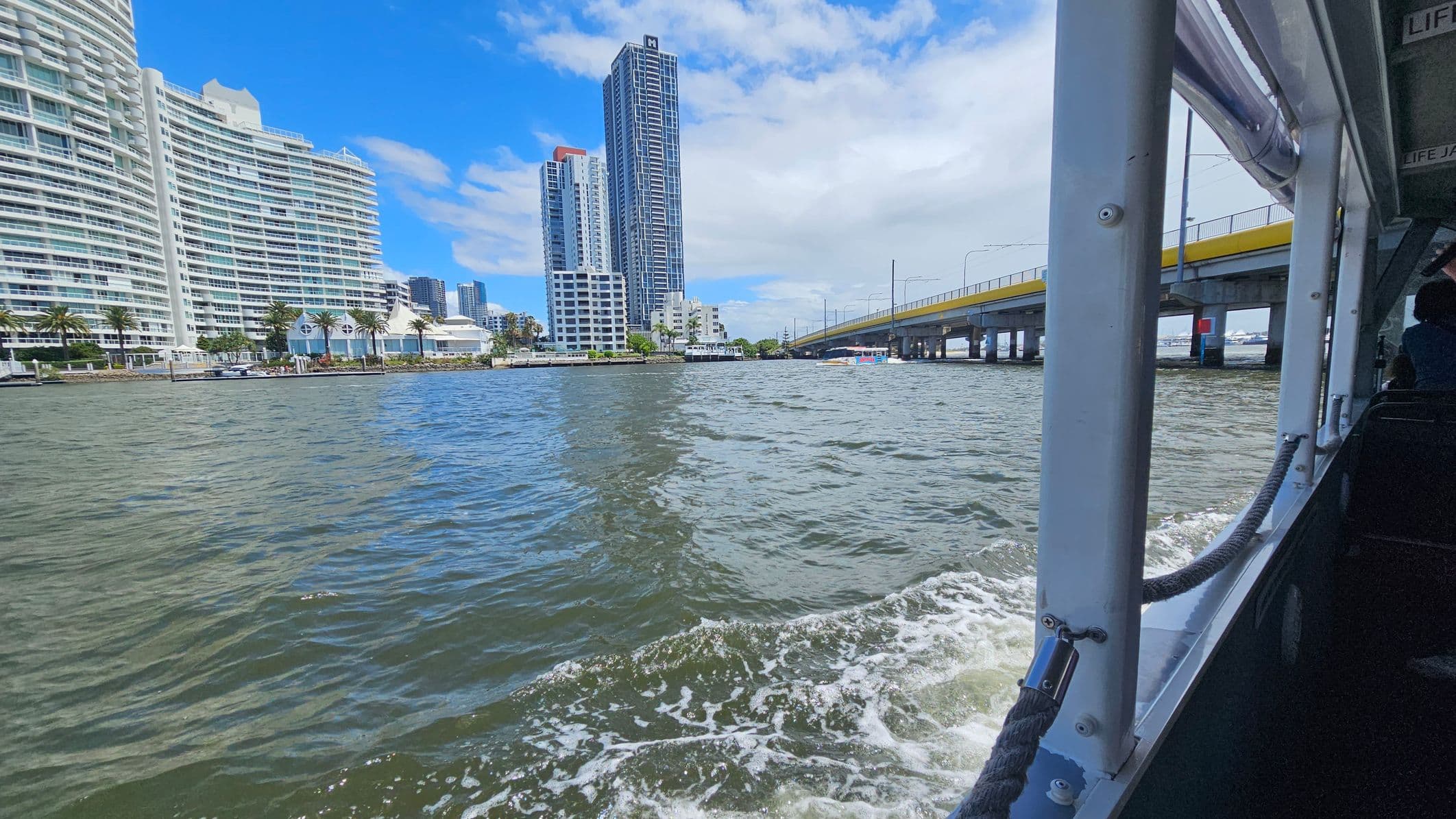 On the left, there are modern high-rise buildings with curved facades and palm trees lining the waterfront. In the middle, a tall skyscraper stands prominently among other buildings. On the right, a bridge with a yellow railing extends across the water. The foreground shows the edge of a boat with a metal railing and a rope, with water splashing against the side. The sky is partly cloudy with patches of blue.
