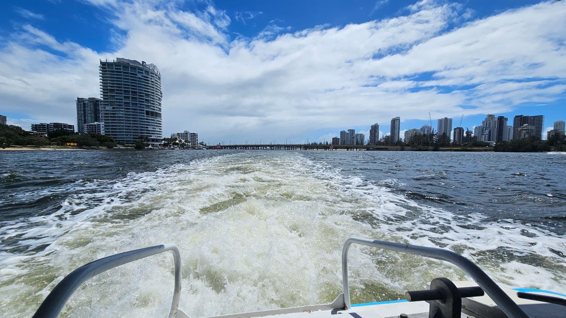 View from a boat looking towards a city skyline. In the foreground, the boat's railing and wake are visible. The left side features a prominent, curved high-rise building near the water's edge, with smaller buildings nearby. On the right, a cluster of tall skyscrapers is visible, with some cranes indicating construction. A bridge spans the water in the distance, connecting the two sides of the cityscape. The water is choppy, reflecting the movement of the boat.