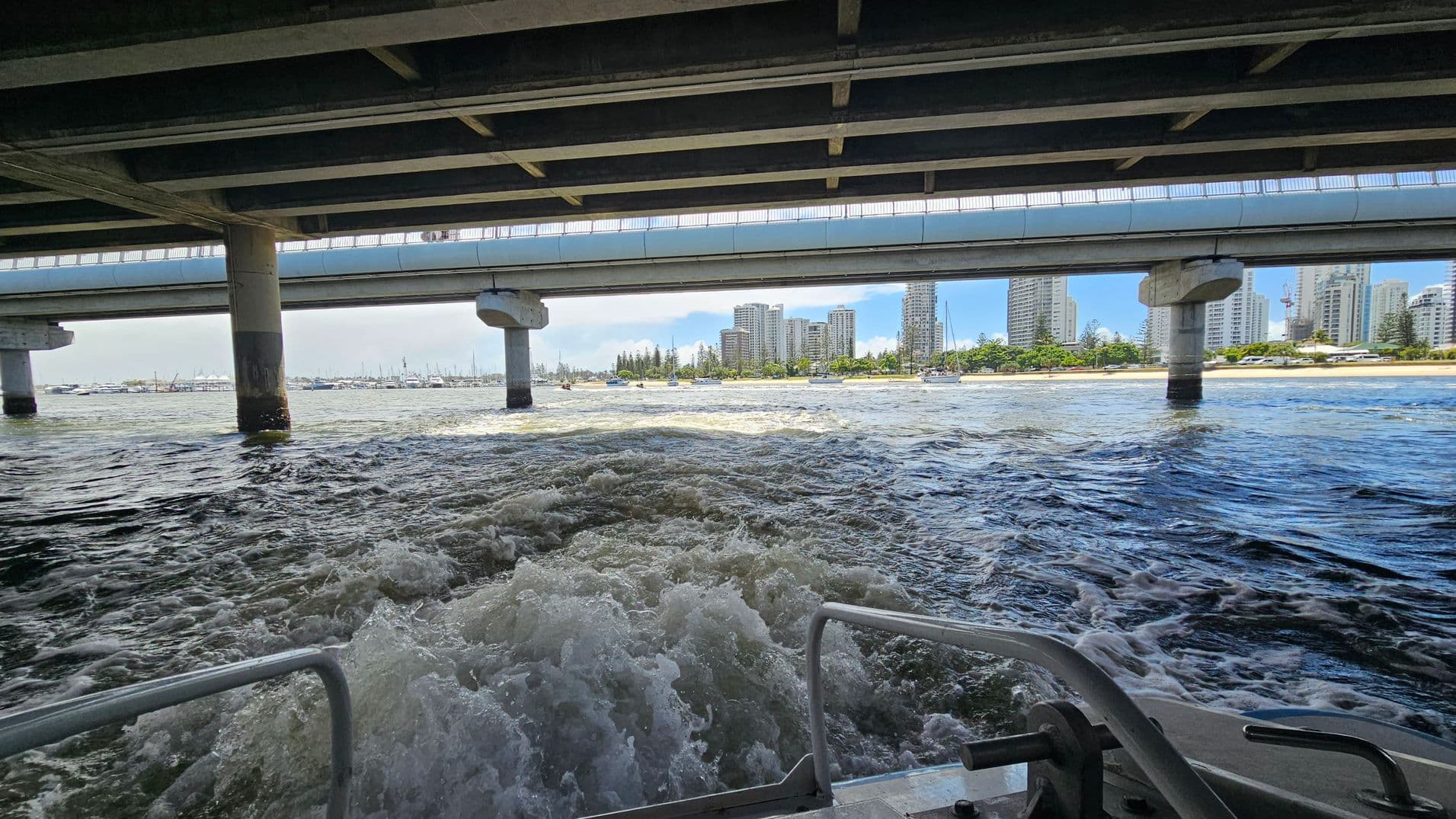Underneath a bridge, with its concrete beams and pillars visible above. In the foreground, churning water is seen from the back of a boat, with metal railings on either side. In the background, a cityscape with tall buildings and a line of trees along the shoreline. Several boats are anchored near the shore, and the water is calm beyond the boat's wake.