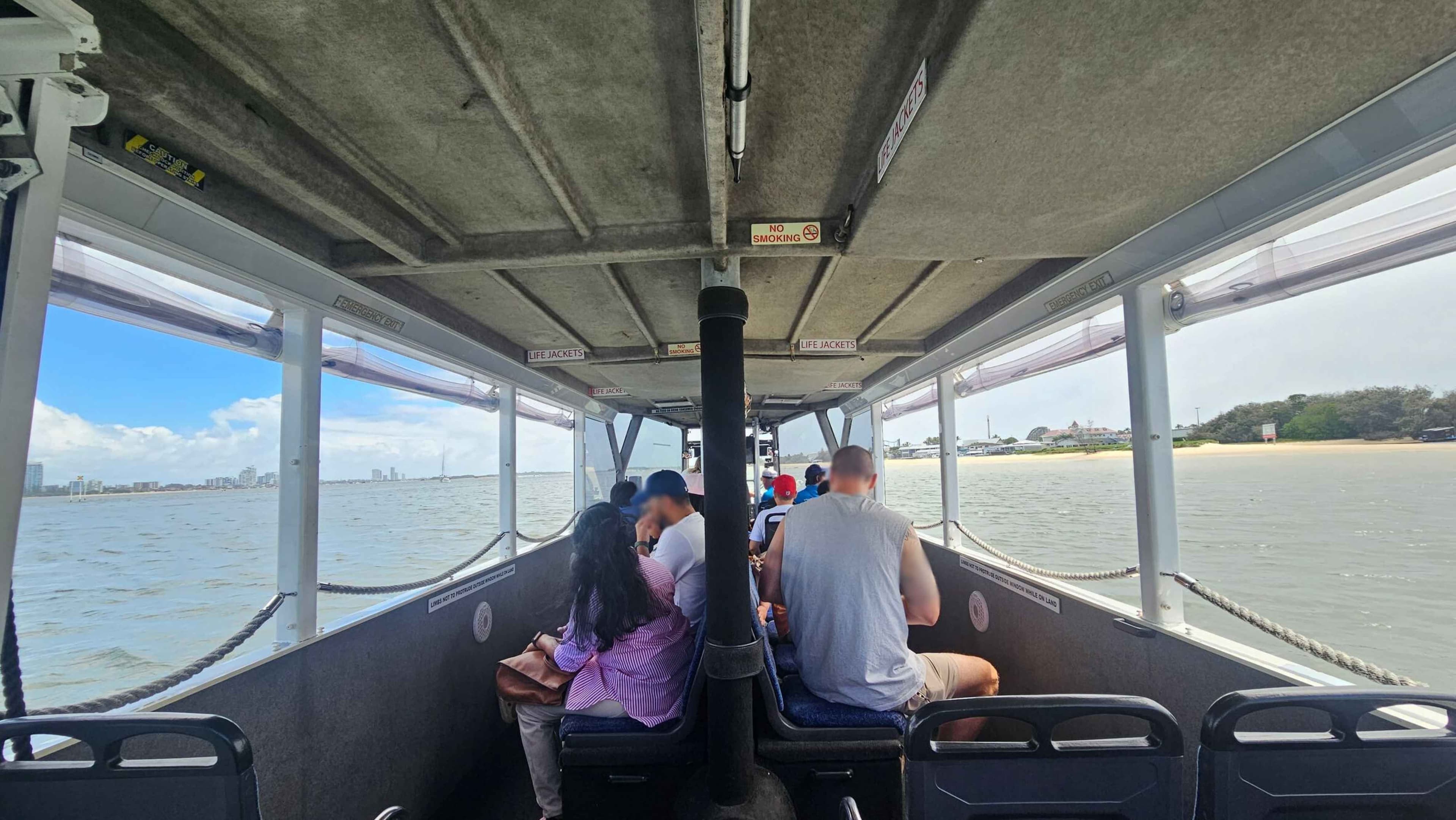 Inside a boat with passengers seated on both sides. The ceiling is green with signs like "NO SMOKING" and "LIFE JACKETS." Large windows offer views of water and distant city buildings on the left, and a sandy shore with greenery on the right. The floor is not visible. The interior has a central aisle with a black column.