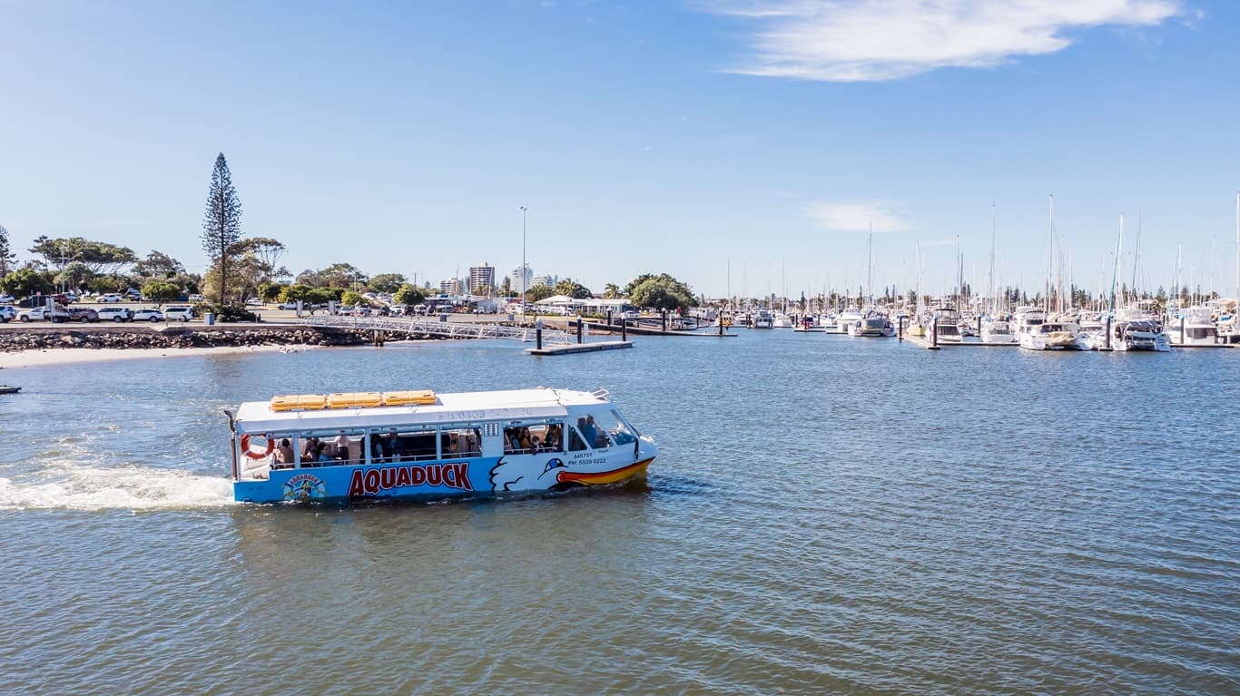 Aquaduck amphibious vehicle in the water. The vessel has orange-white color schemes, blue waves, a logo, and the word “AQUADUCK” on the side. The front resembles the head of a smiling duck with a big yellow beak. Found on the end of the vessel are two orange life buoys. The passengers sitting in their seats are feasting their eyes on the beautiful view of the surroundings and the calm waters of the Mooloolaba River. The background is a marina with many boats and yachts.