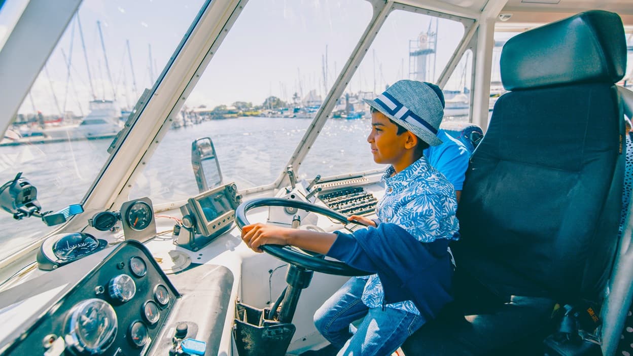 A boy sitting in the captain’s black chair of the Aquaduck is assisted by staff wearing a blue polo shirt. The boy is wearing a blue and white Hawaiian shirt and a grey baseball hat with white and blue accents. The boy is holding the black steering wheel while staring directly in front. Lots of navigation controls and gauges are visible on the vessel's dashboard. Other yachts and boats can be seen on the glass window.