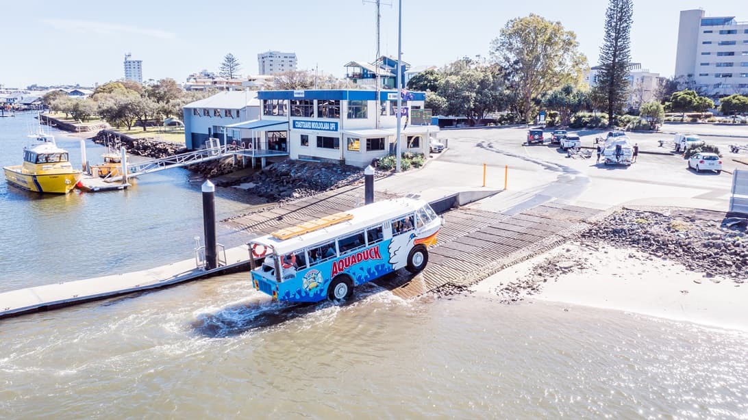 The Aquaduck, leaving the water and ascending to a steep boat ramp that has horizontal lines. There is a building to the left with white and blue color schemes. It has a bridge with metal handrails that connects to a boat dock. A yellow boat can be seen on the dock.  The park with trees, the parking area, and the pathway leading to the main facility can be identified from this image.