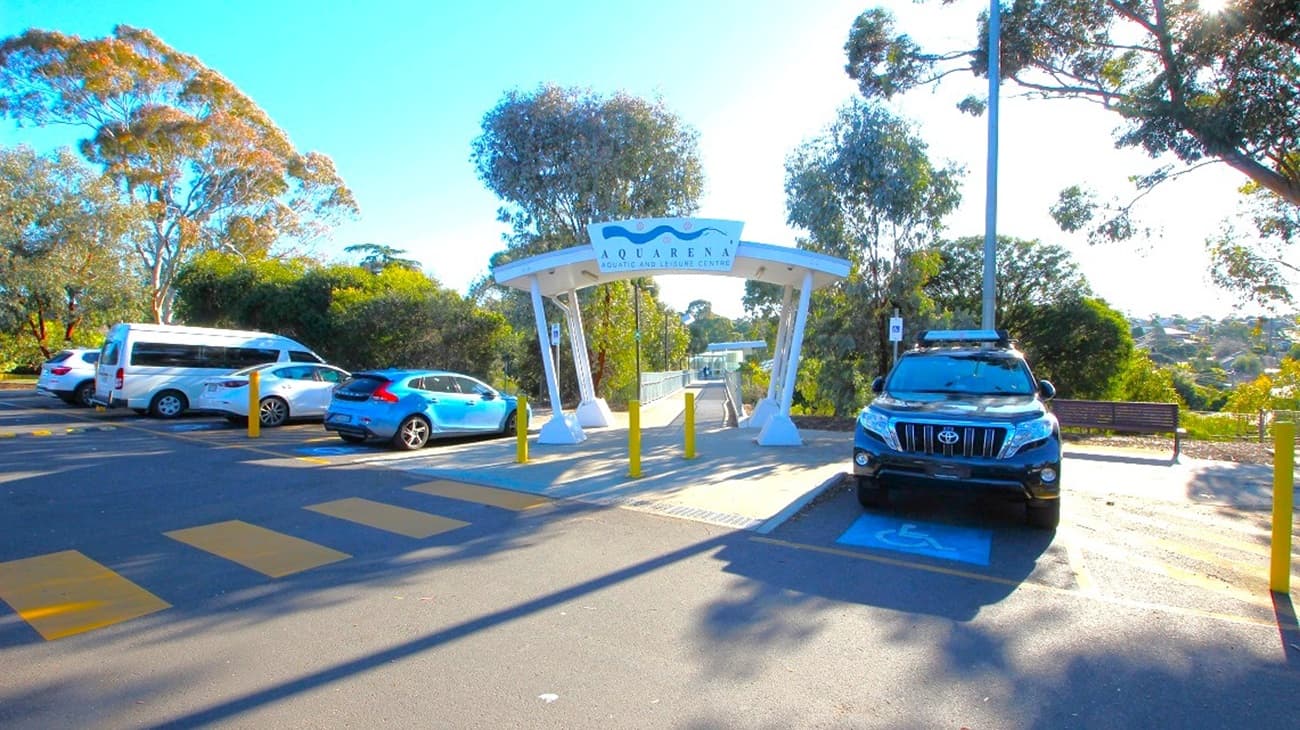 A parking area with several vehicles, including a white van and a blue car on the left, and a black SUV on the right. In the centre, a white archway with a sign reading "Quarry" leads to a pathway. Yellow bollards are in front of the archway. Trees and greenery surround the area, with a bench visible on the right. The ground is marked with yellow pedestrian lines, and a tall streetlight is on the right.