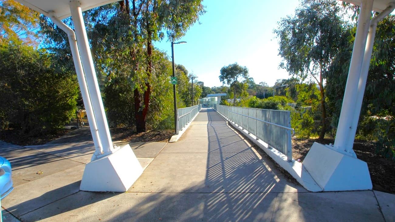 A walkway with a metal railing on both sides extends into the distance, flanked by tall trees on the left and right. The path is paved and slightly elevated, with shadows cast by the railings. In the foreground, there are two large white support structures on either side. A blue car is partially visible on the left. The background features more trees and a clear sky.