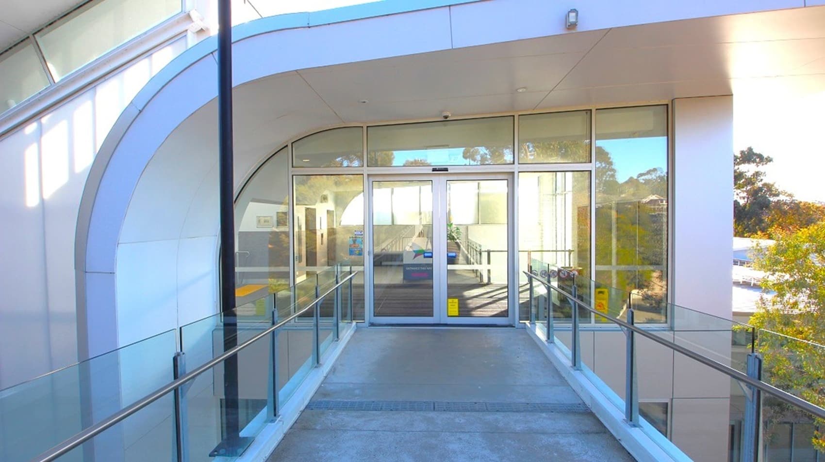 A modern entrance with glass double doors in the centre, surrounded by large glass panels. The structure has a curved design with a white exterior. A concrete ramp with metal railings leads up to the doors. On the right, trees are visible through the glass. The ceiling is smooth and white, with a light fixture above the doors.