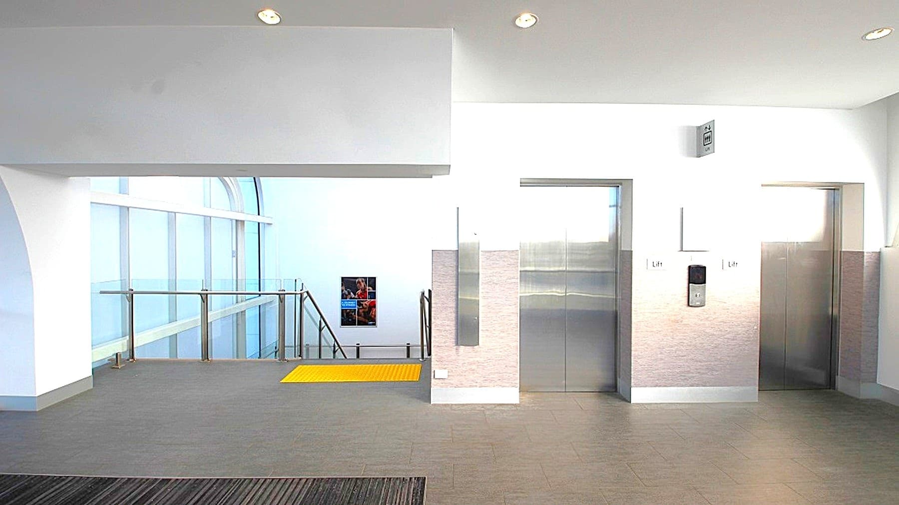 A modern interior space with two lift doors on the right, featuring metallic finishes. To the left, a staircase with a glass railing descends, and a yellow safety strip is visible on the floor near the stairs. The walls are white, and the ceiling has recessed lighting. The floor is grey, complementing the minimalist design. A small window is visible in the background, providing natural light.