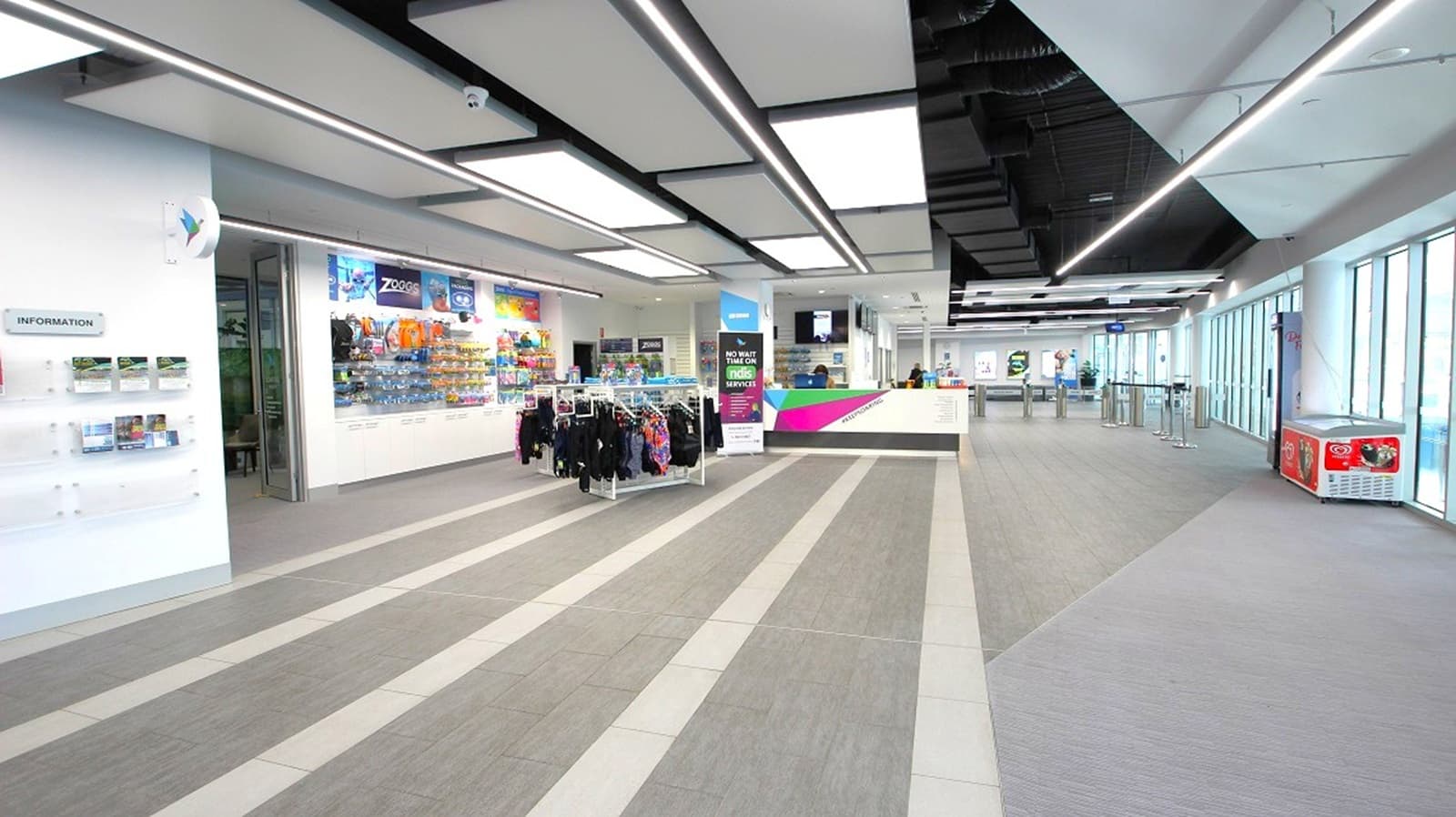 A spacious, modern interior with a grey-striped floor and a ceiling featuring rectangular light panels. On the left, there is a wall with information displays and a shop area with racks of clothing and shelves of merchandise. In the centre, a digital sign stands near a counter. The right side has large windows allowing natural light, and a small vending machine is visible. The area is open and well-lit, with a sleek, contemporary design.