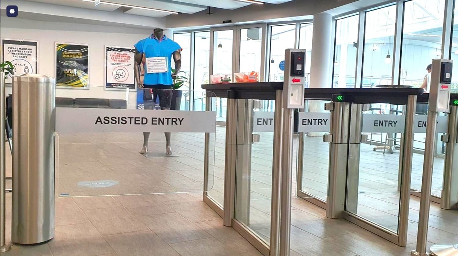 A modern entry security checkpoint with glass doors labeled “ENTRY” and a barrier marked “ASSISTED ENTRY.” A mannequin or person stands inside the entrance area, and turnstiles/automatic doors are visible.