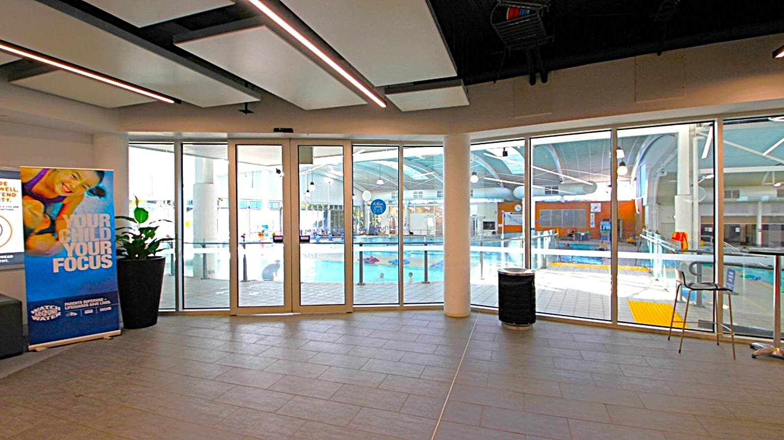 A spacious indoor area with large glass doors and windows in the centre, providing a view of a swimming pool area with clear water and poolside equipment. On the left, there's a promotional banner next to a potted plant. On the right, a tall chair and a small table are positioned near the windows. The ceiling features modern lighting fixtures, and the floor is tiled in a dark colour.
