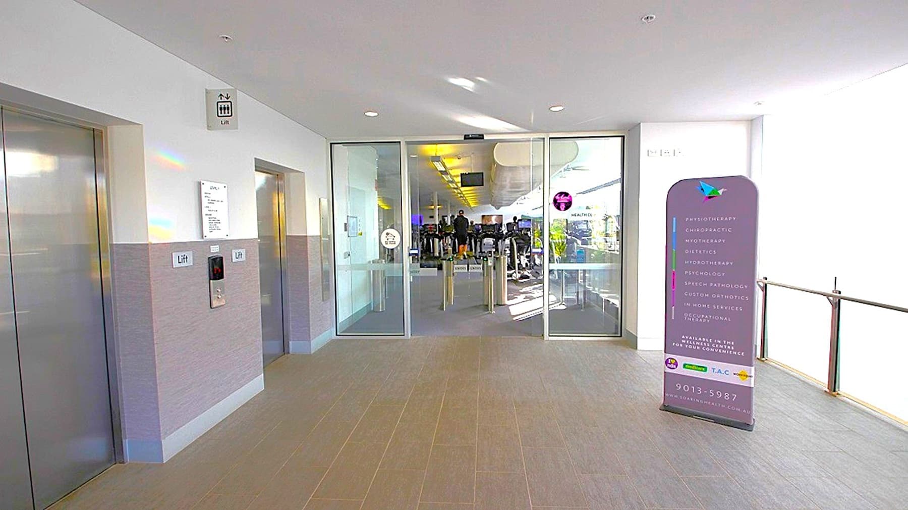 On the left, there are two lift doors with a sign above indicating their location. In the centre, glass doors lead into a gym area with exercise equipment visible inside. On the right, a purple sign with text stands near the entrance. The floor is tiled, and the ceiling is white with recessed lighting.
