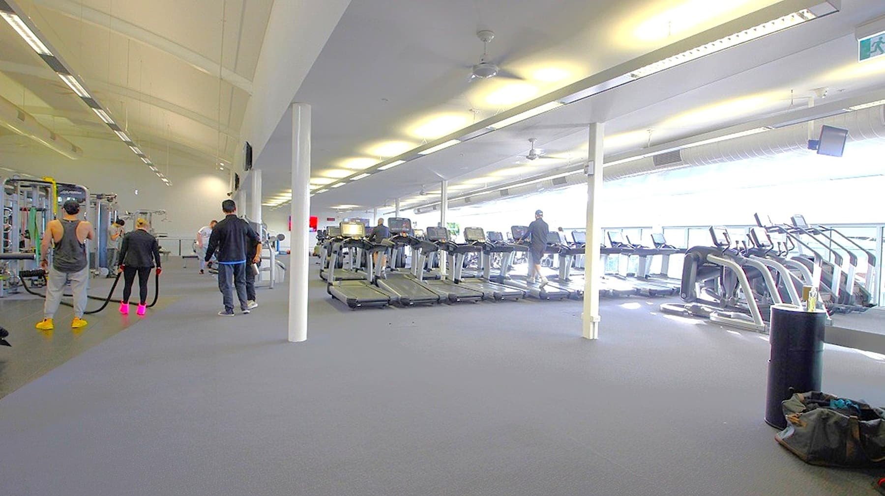 A spacious gym interior with a grey carpeted floor. On the left, several people are using exercise equipment, including a person holding battle ropes. In the centre, a row of treadmills is visible, with a few individuals using them. On the right, more cardio machines are lined up near large windows that let in natural light. The ceiling is high with fluorescent lights, and a white pillar is in the middle of the room. A black cylindrical object and a bag are on the floor in the foreground.