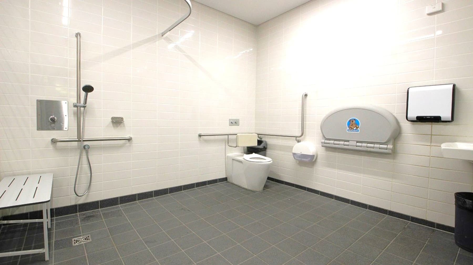 A spacious, accessible bathroom with light-coloured tiled walls and a grey tiled floor. On the left, there is a shower area with a handheld showerhead and a metal grab bar. A bench is positioned nearby. On the right, a toilet with support rails is visible, along with a baby changing station mounted on the wall. A paper towel dispenser and a waste bin are also present. The ceiling is plain and light-coloured.