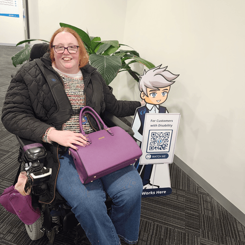 A woman in a wheelchair sits beside a Cérge cardboard cartoon cutout with a QR sign.