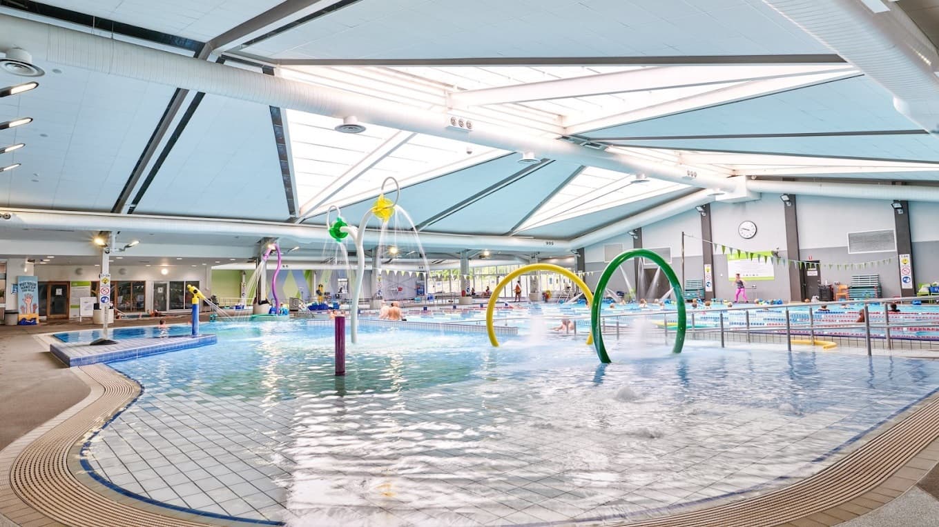 An indoor swimming pool features colourful water play structures on the left, including arches and hoops that spray water. On the right, designated swimming lanes are outlined by lane dividers. The foreground displays a children's splash area with a beach entry. In the background, there is a pool deck with a seating area and a large clock on the wall. Another pool with a ramp entry is situated to the right. The ceiling is equipped with skylights, large air conditioning vents, and light fixtures.