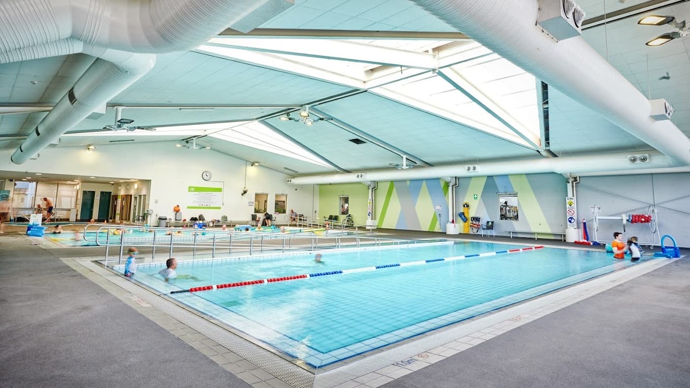 Two indoor swimming pools, each equipped with a ramp and handrails and currently occupied by swimmers, adjoin a third pool sectioned by floating rope dividers. The walls are painted green and blue with X-shaped patterns, and the ceiling features exposed metal beams, skylights, large air conditioning vents, and light fixtures. Posters and banners are affixed to the walls. A glass wall at the far end secures the aquatic facility. Doors leading to the changing rooms are visible at this end.