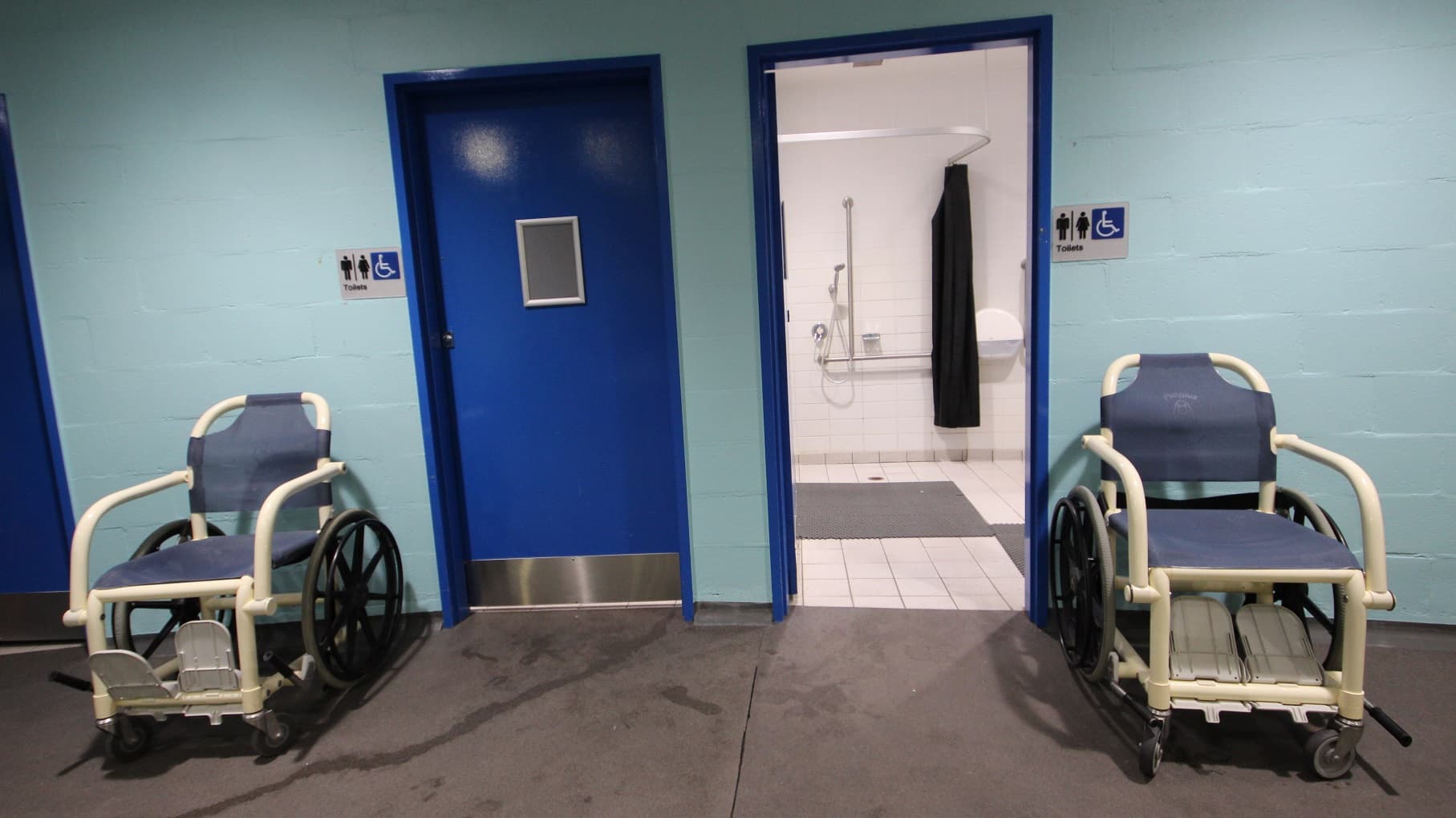 Two manual wheelchairs flank a central corridor: on the left, a closed blue door with a number, and on the right, an open door with leading into a bathroom with visible safety handrails and a shower area. Accessibility and unisex toilet signages are affixed to the wall. The walls are painted turquoise, and the floor is a grey non-slip surface.
Posters and banners are affixed to the walls.