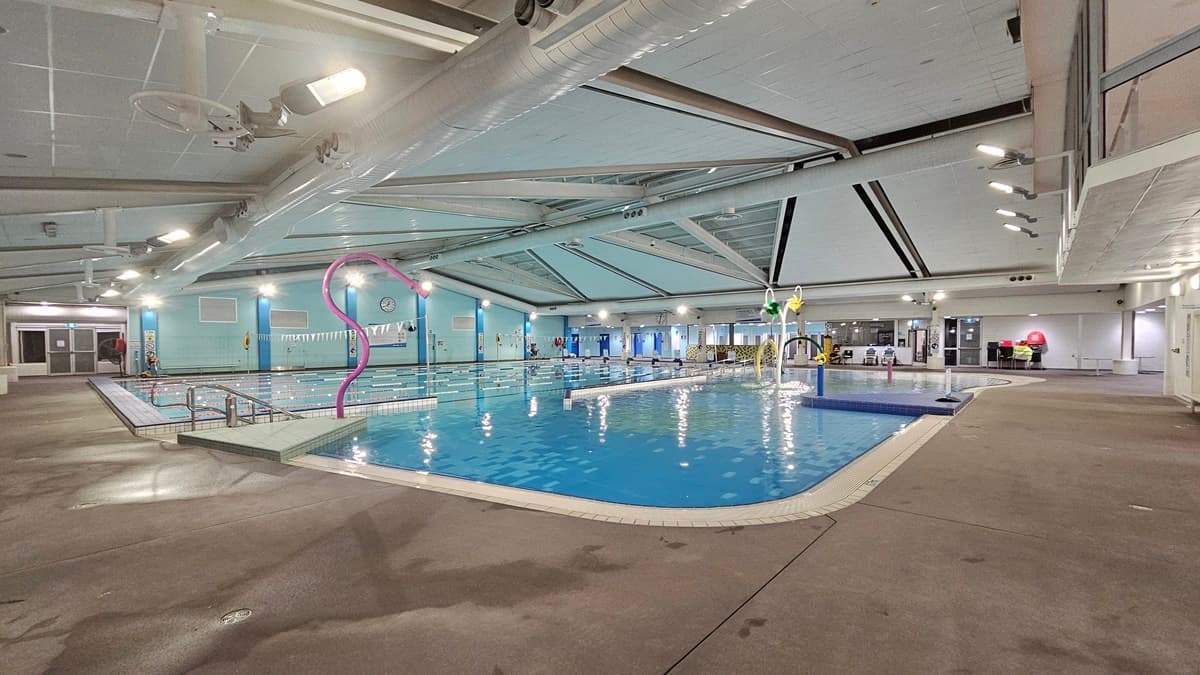 An indoor swimming facility features steps with handrails leading into a pool adorned with colourful hoops, sprayers, and a fountain. Adjacent to this is a larger pool bordered by floating ropes and triangular banners. The ceiling is equipped with large air conditioning vents, metal beams, and skylights. A glass wall provides views of the outside. Reminder posters are visible in the area. On the far right, chairs are stacked on the pool deck.