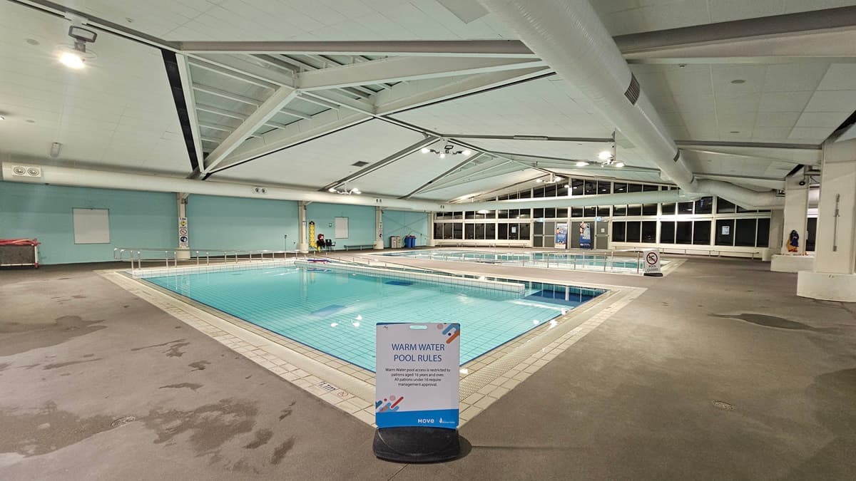 Two pools, each equipped with a ramp and handrails, are separated by a pool deck. A freestanding banner is positioned at the corner of the pool. The wall on the left is constructed from concrete and painted turquoise, while the central wall is made of glass. The ceiling features large air conditioning vents, metal beams, and skylights. Large banners and reminder signs are visible around the area.