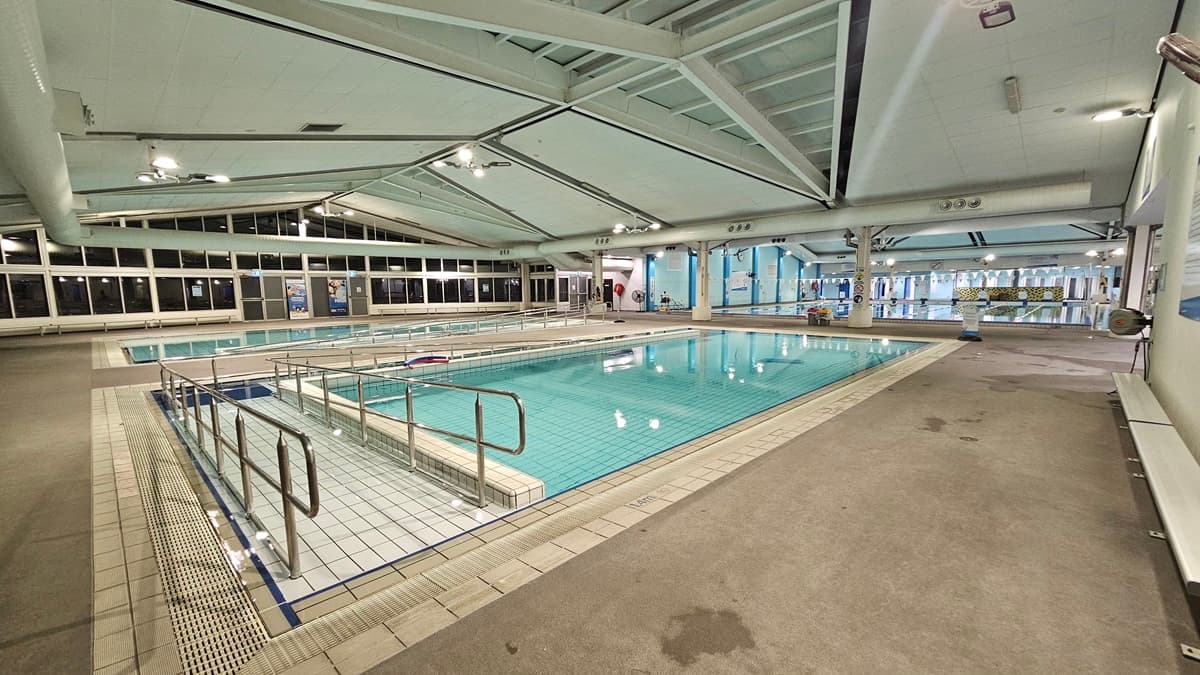 An indoor pool with a ramp and handrails is adjacent to another pool, also equipped with handrails. On the right, a bench chair is positioned on the pool deck against the wall, and on the left, bench chairs are arranged against the glass wall. Large posters, a fire hose reel, and a fire extinguisher are affixed to the wall. At the far end, a children's pool is visible.
