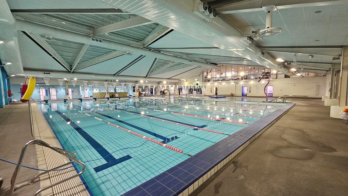 An indoor pool features a ramp with handrails and colourful floating ropes, with triangle banners hanging across it. Adjacent to this is another pool, also equipped with handrails, and next to it, a children's pool with steps and handrails. A life-size surfboard decoration is positioned in the left corner. At the far end, doors leading to the changing facilities are visible. The ceiling is outfitted with large air conditioning vents, metal beams, and skylights.