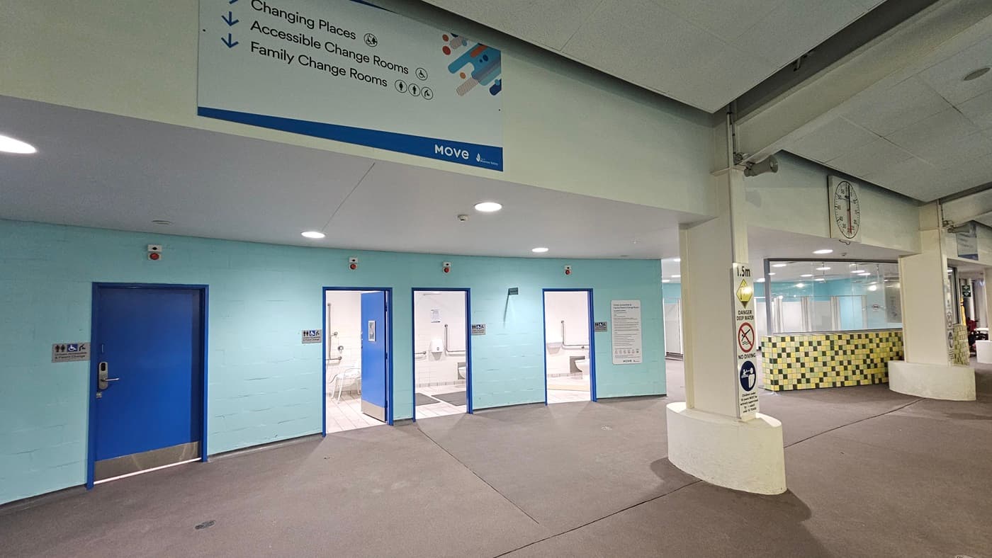 An indoor facility features a turquoise wall on the left with four blue doors, each labelled with gender access signs and signage for changing places and family rooms. On the right, there is another changing room with multiple cubicles, characterised by mosaic and glass walls. Large directional posters and a large round clock are affixed to the upper wall. Additional safety reminder posters are attached to the columns.