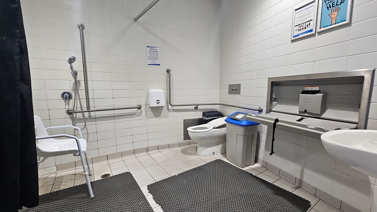 The accessible bathroom features a toilet with handrails on the right and a shower with hand grabs on the left. A white plastic chair is positioned against the left wall. To the right, there is a change table, a wash basin, a rubbish bin, and a tissue dispenser. The floor is white, equipped with black non-slip mats, and the walls are white with posters affixed.