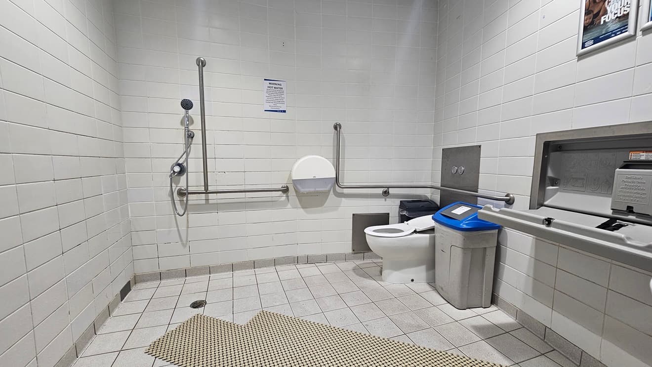 The accessible bathroom includes a toilet with handrails on the right and a shower with hand grabs on the left. A tissue dispenser and a rubbish bin are positioned against the wall. To the right, there is a change table. The floor is white with grey non-slip mats, and the walls are white with posters affixed.