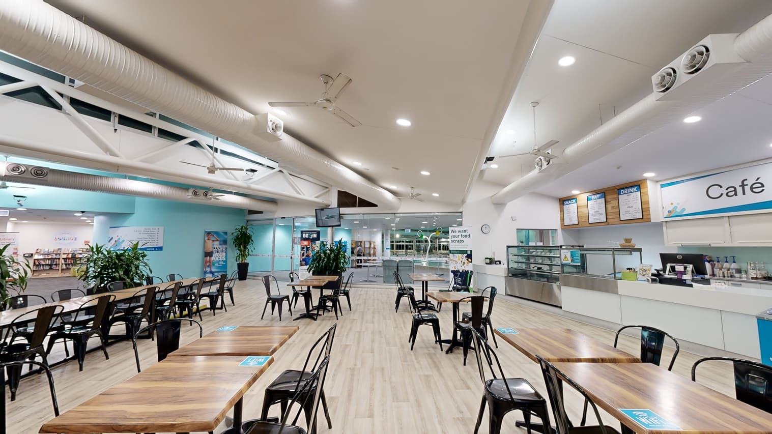 Rows of movable tables and black chairs are arranged in the centre. To the right, there is a cafe counter with a food display rack, above which the menu is posted on the wall. Indoor plants in large planter boxes are placed along the sides. The floor is timber, and the ceiling is white, featuring large air conditioning vents, ceiling fans, and lights. In the background, a shelf filled with books is visible. Posters are affixed to the wall.