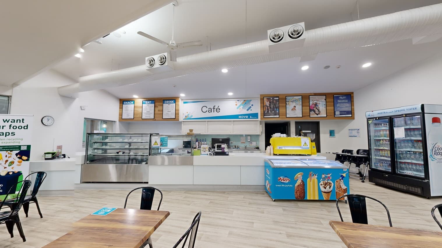In the centre is a cafe counter with a white facade. The cafe's menu and an advert poster are affixed to the timber wall. A food rack is positioned next to the cashier area. To the left, a fridge filled with cold drinks stands next to an ice cream freezer. Tables and chairs are arranged in front, with a retractable banner and a clock positioned to the left. The flooring is timber, and the white ceiling is equipped with lights, large air conditioning vents, and ceiling fans.