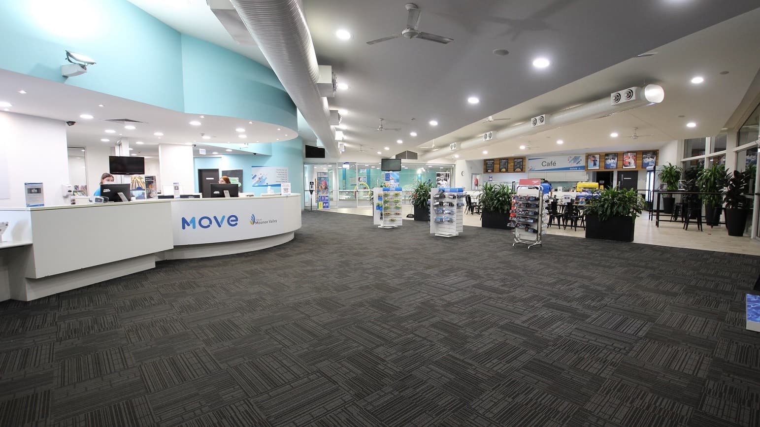 To the left, there is a reception desk with a white front, topped with computers and brochures. Next to it, rows of display racks hold goggles and swimming accessories. Further left, a glass door offers a view of the car park outside. Large planter boxes with indoor plants are placed around the area. The ceiling is white, fitted with light fixtures, posters, ceiling fans, and large air conditioning vents, which complement the light blue hues of the walls. The floor is covered with a grey carpet.
