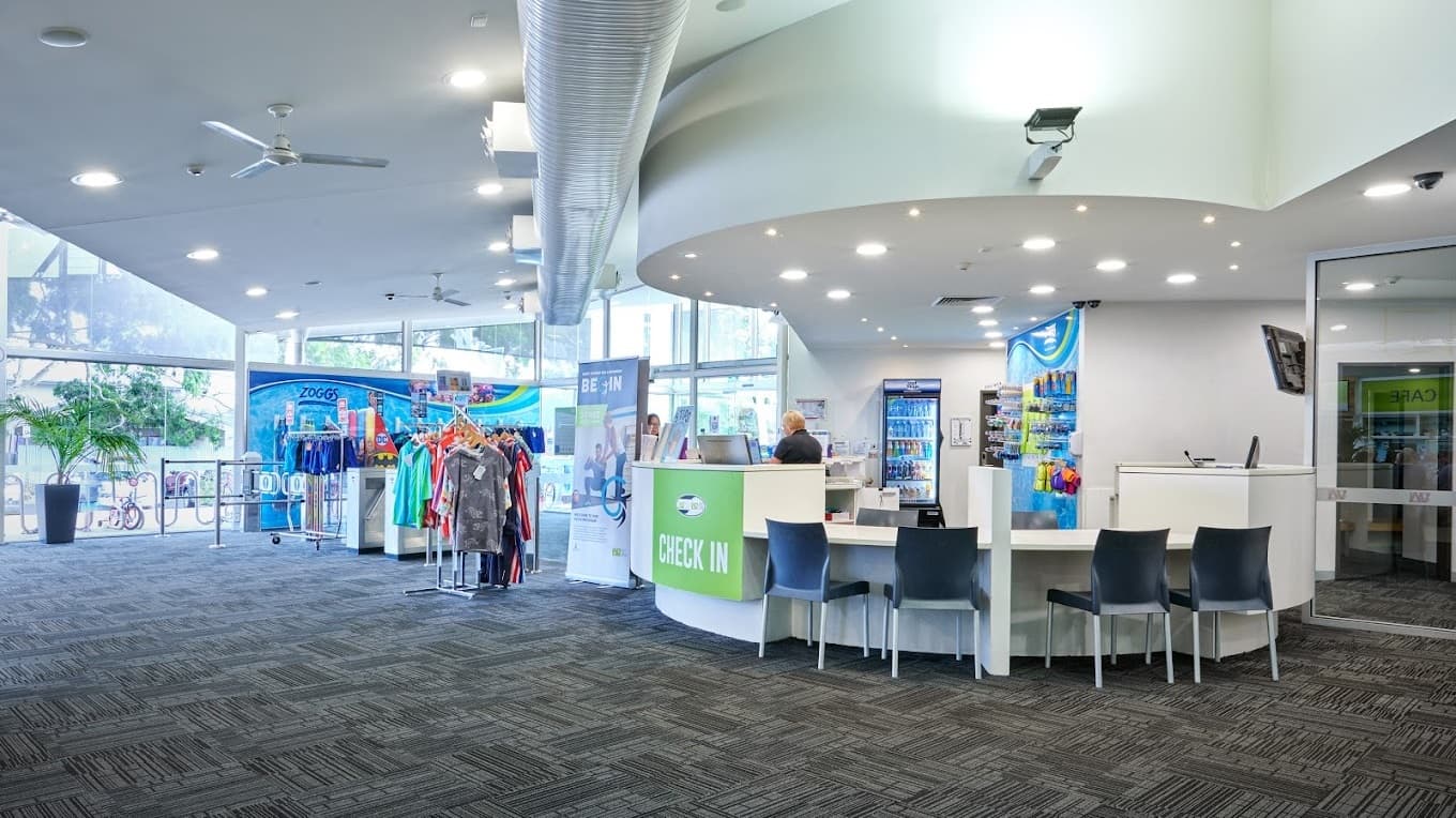 The check-in counter features a concave design with a green "Check In" banner. Black chairs are tucked under the desk. Behind, a fridge with beverages is placed next to a display rack showcasing goggles. To the left, a retail area displays clothing and swimwear, adjacent to a large retractable advert banner. The glass walls are adorned with a large advert banner. The white ceiling is fitted with light fixtures, ceiling fans, and air conditioning vents. The floor is covered with grey carpet.