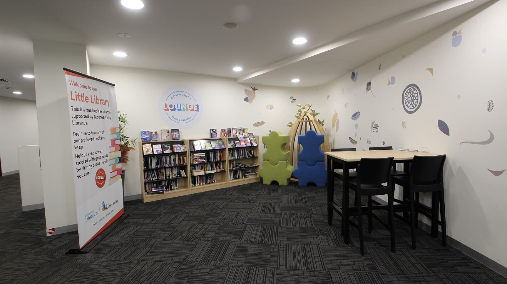 The community lounge features shelves against the wall, displaying pre-loved books. Next to this is a life-sized puzzle piece decoration and wooden plant decor. To the right, there is a tall table accompanied by high chairs. A large retractable banner is positioned on the left, adjacent to an indoor plant. The walls are white, adorned with pastel prints, and the floor is laid with grey tiles.