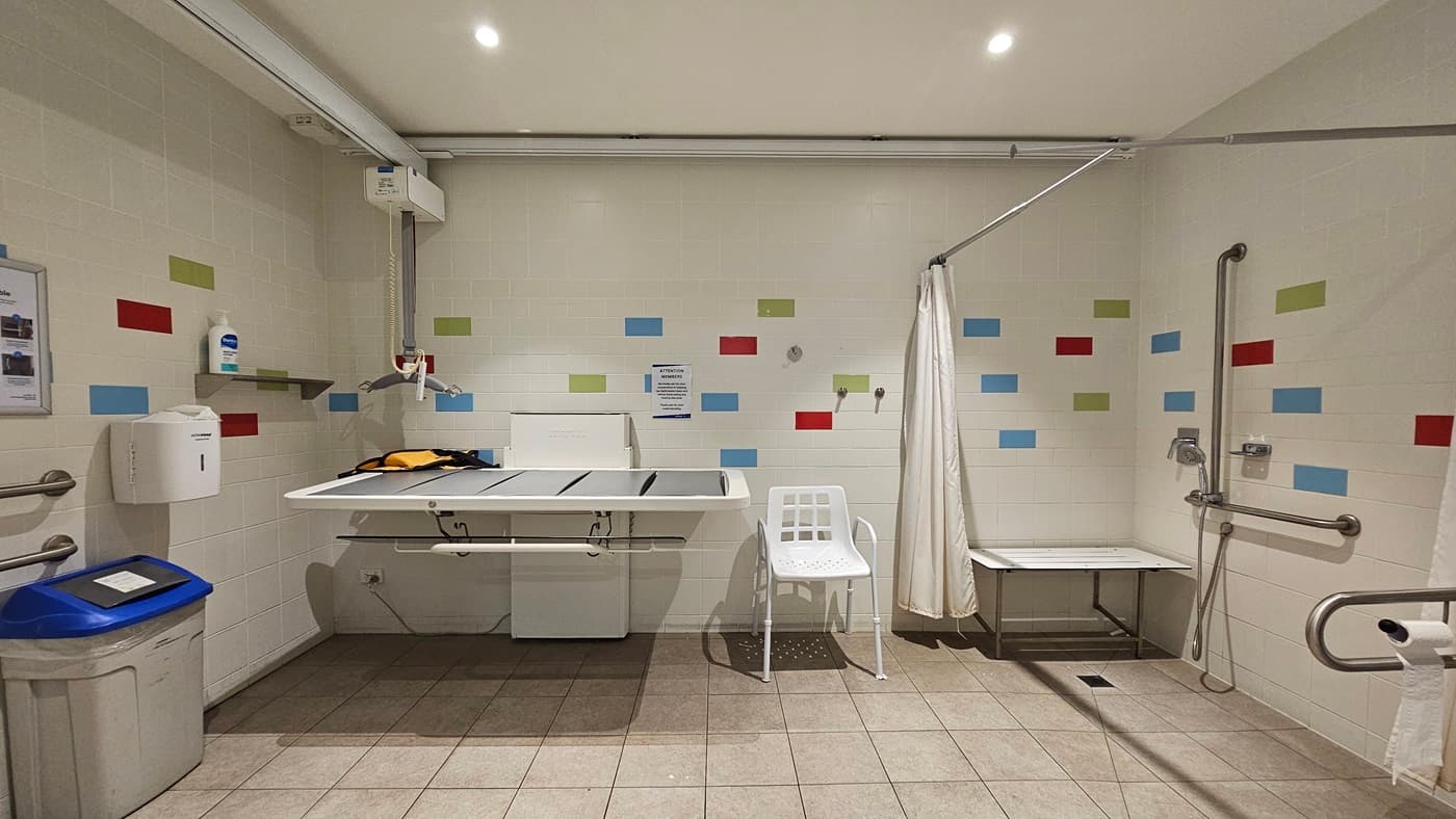 The Changing Place features a life-sized changing table, a chair, and a bench mounted to the wall. To the right, there is a shower equipped with a curtain and hand grabs. To the left, a hoist, liquid soap dispenser, rubbish bin, tissue dispenser, and hand grabs are present. The walls are white with an accent of multicoloured tiles, and the floor is covered with light brown tiles. A poster is affixed to the wall, and the area is well-lit, illuminated by LED lights.