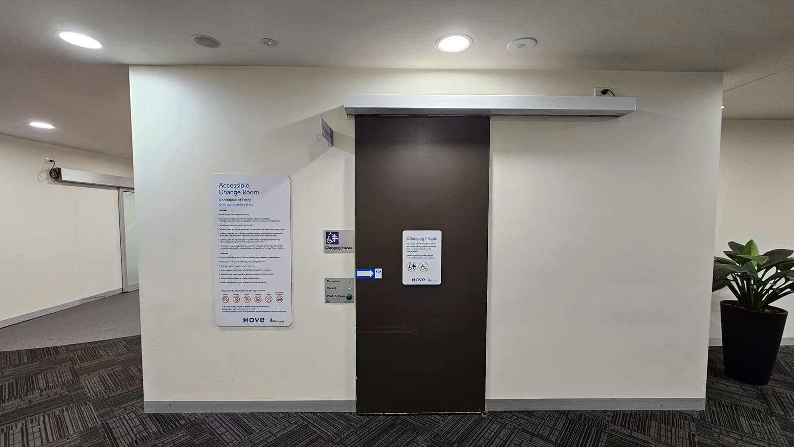 A brown door leads to the accessible change room. Affixed to the wall next to it is a poster with reminders, gender access signage, and an access button. To the left of the door is a lobby, and to the right, there is an indoor plant in a large pot. The walls and ceiling are white, fitted with light fixtures, while the floor is covered with grey carpet.