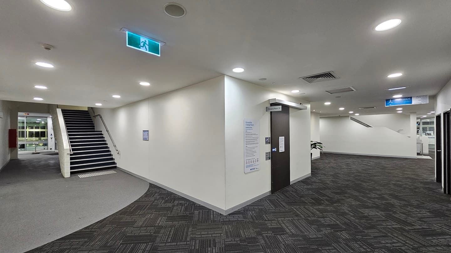To the right, a brown door leads to the accessible change room, with a poster displaying reminders, gender access signage, and an access button affixed next to it. To the left of the door is a lobby, and on the right, an indoor plant stands in a large pot. The walls and ceiling are white, equipped with light fixtures, and the floor is covered with grey carpet. On the left, stairs fitted with handrails and truncated domes are present. Behind the stairs, the aquatic facility is visible.