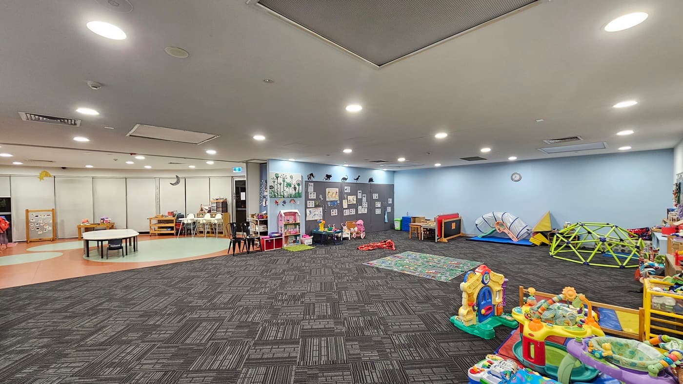 An interior view of a well-lit room features patterned grey carpet flooring. To the left, colourful children's artwork adorns the walls above shelving filled with books, alongside a play area with toys. To the right, there is a pram and additional toy storage. Shelves displaying toys are arranged against the walls. A colourful map carpet is positioned in the middle of the room. The ceiling is white, and equipped with recessed lighting and air conditioning vents.