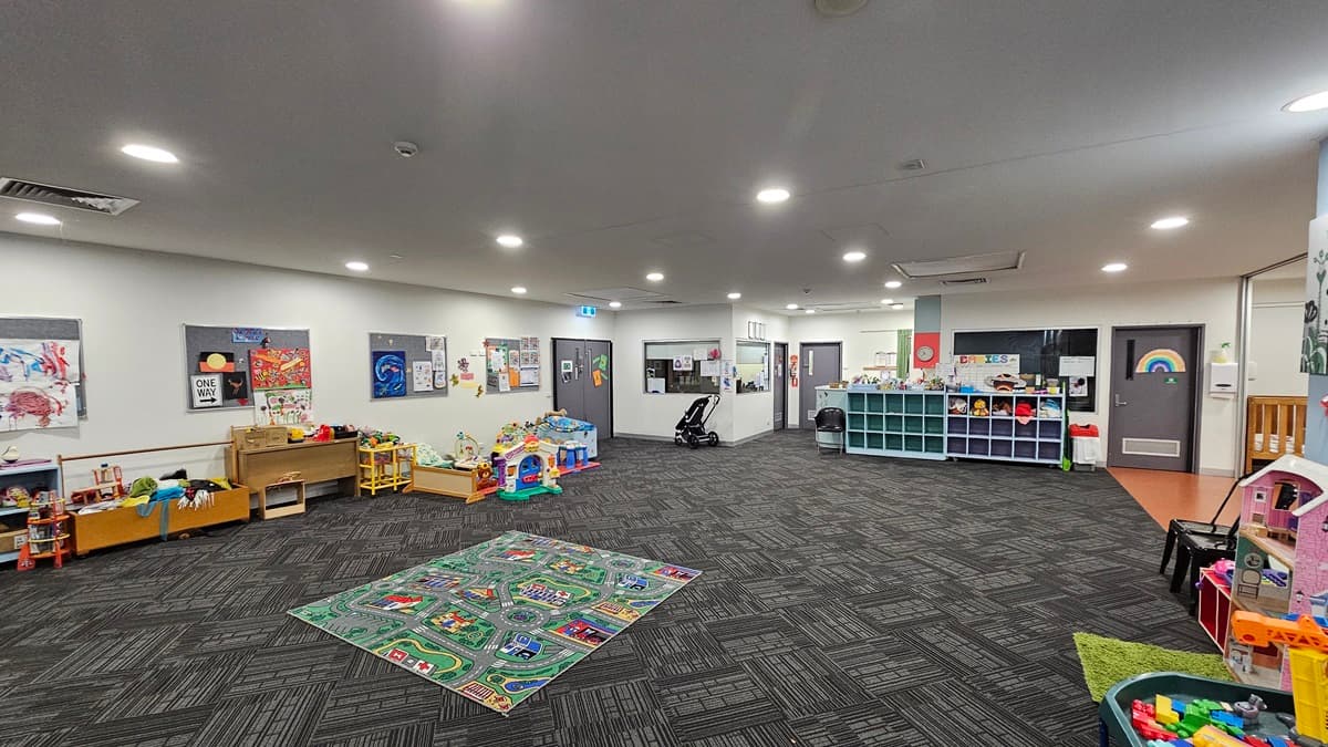 The interior of a spacious room features a patterned grey carpet and a plain ceiling. On the left, there are tables with chairs and a shelving unit, while the right side is filled with various children's toys and play structures. In the background, there is a notice board, and the foreground is scattered with more toys. The wall has light colors adorned with posters