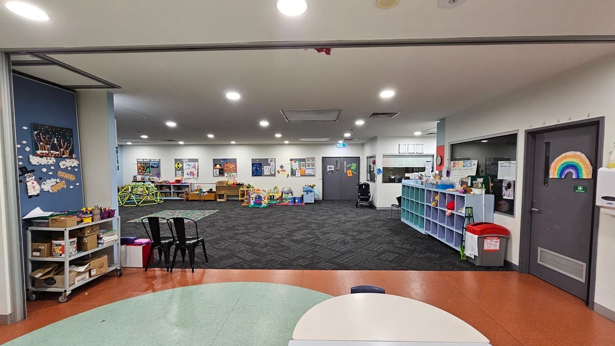 A spacious indoor area with a patterned grey carpet. To the left, a blue wall with children's artwork, shelving with toys, and a small table with chairs. On the right, a white wall and door with a rainbow motif. The foreground shows a white round table edge, while the ceiling is white with recessed lighting.
