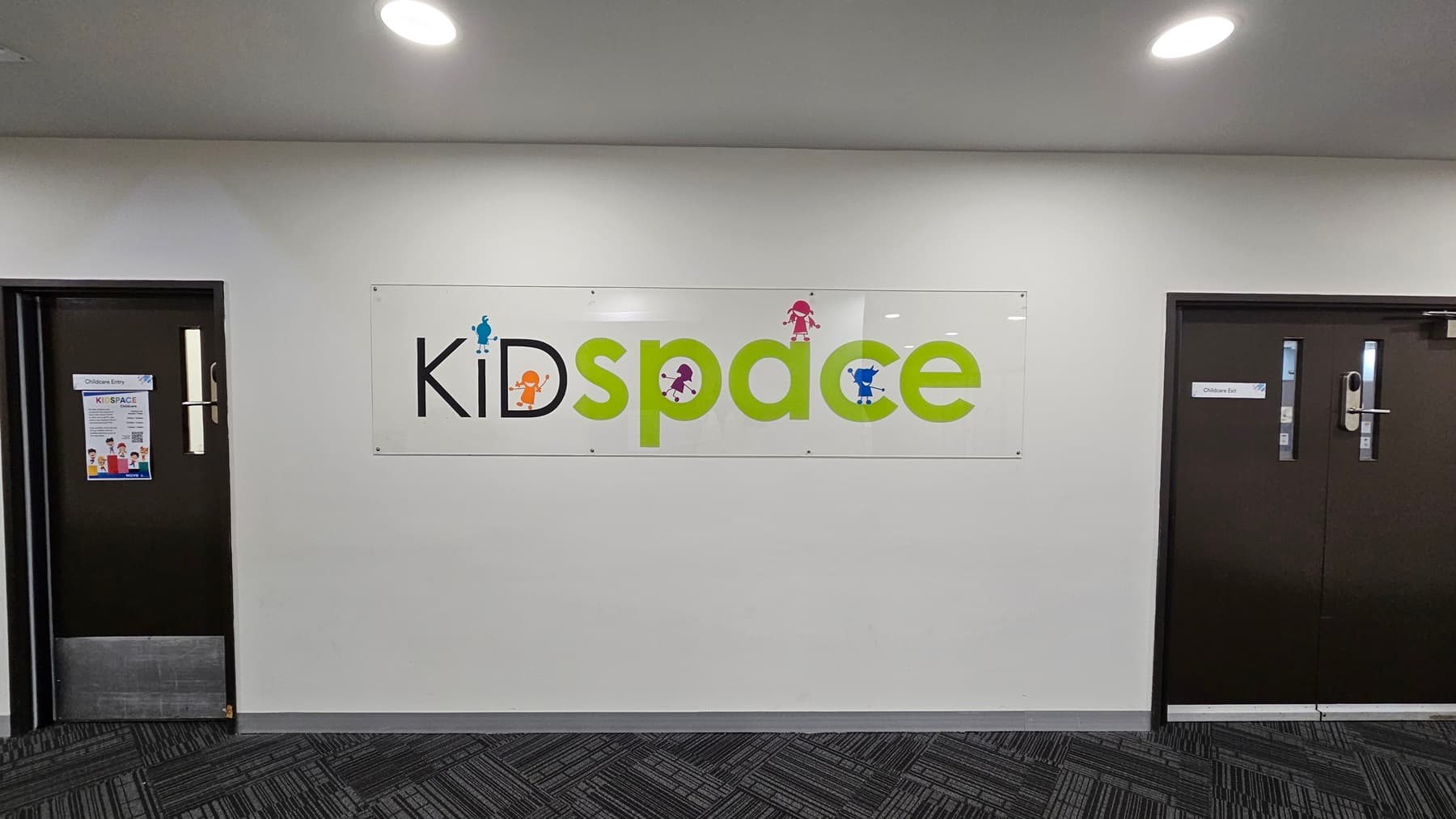 A sign reading "Kidspace" in colourful letters is centrally mounted on a light grey wall, flanked by two closed dark doors with metal handles, one on each side. Flyers are attached next to the left door. The ceiling and floor are not visible.