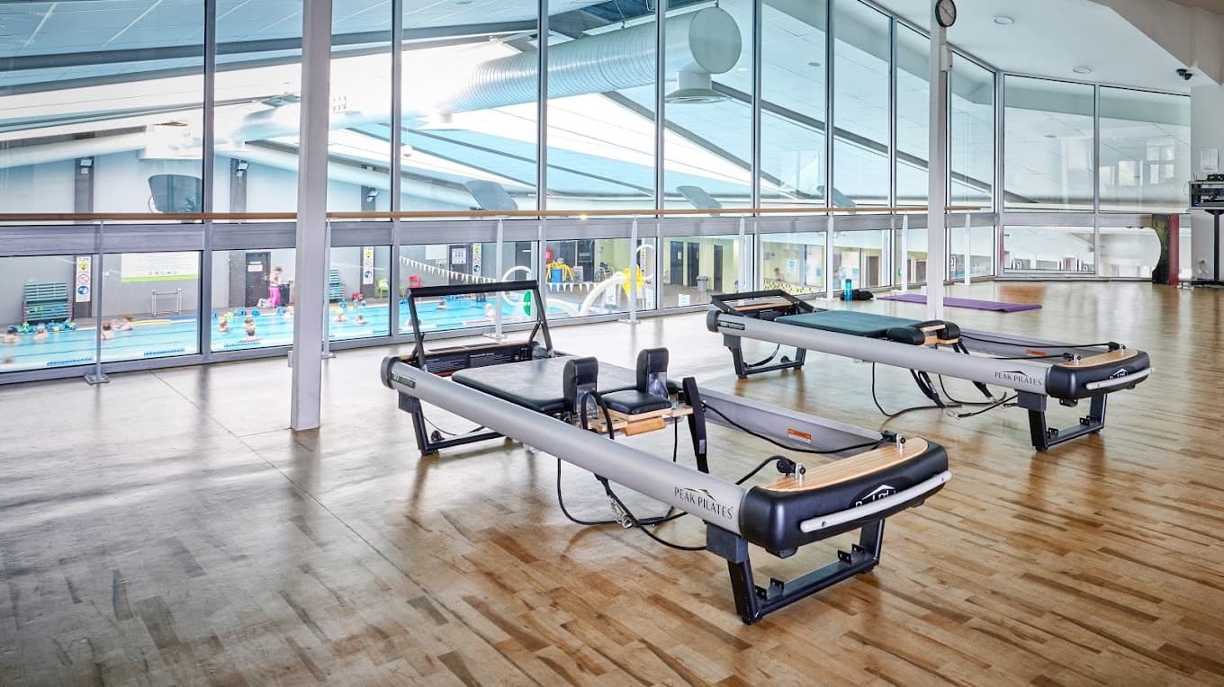Interior of a well-lit space with timber flooring overlooking the aquatic facility below.  Two black and silver pilates reformer machines occupy the foreground, centered in the room. Overhead, a modern ceiling with circular lights and visible ductwork extends throughout the area.