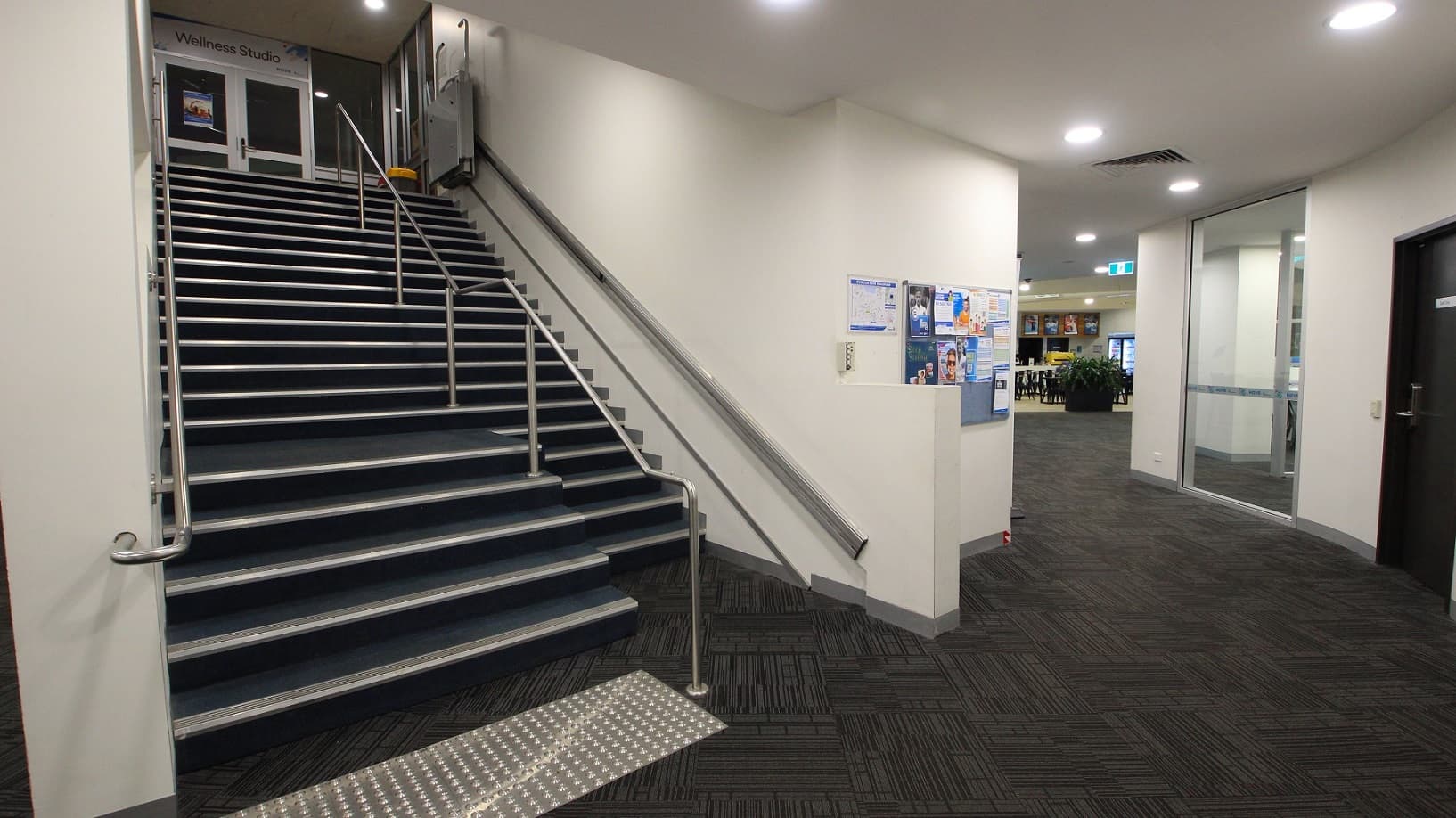 A corridor features a staircase on the left, equipped with handrails and truncated domes, leading to an upper level. A handrail runs parallel to the steps. On the right, a glass door opens into a room, next to another black door with a handle. The floor is patterned with dark geometric shapes, and the ceiling is fitted with recessed lighting. A noticeboard hangs on the back wall. In the background, a café is visible.