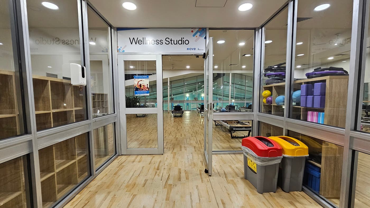 An interior view of a wellness studio features outward-opening glass doors with a poster in the foreground. To the right, exercise balls and mats are neatly stacked on shelves, and a recycling bin is positioned before the door entrance. On the left, a wooden display rack is placed. The flooring is made of timber, and above, a drop ceiling with recessed lighting can be seen.