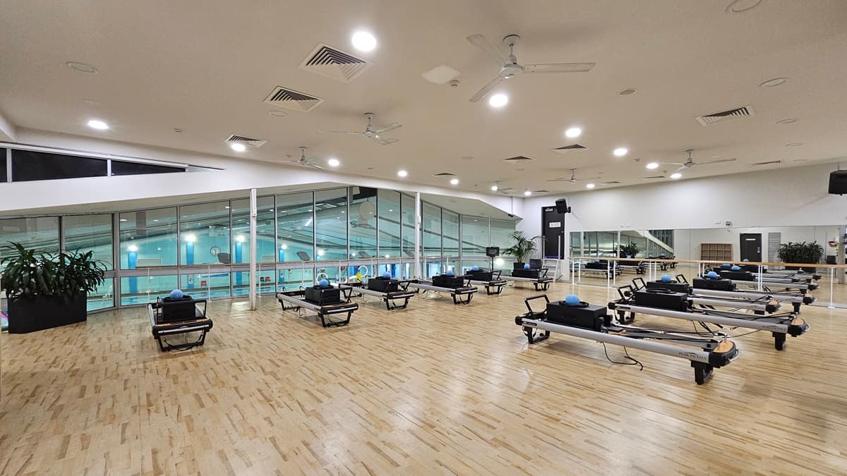 A spacious, well-lit room features a timber floor and a white ceiling equipped with multiple fans, recessed lights, and air conditioning. To the left, large windows offer a view of the aquatic facility below. To the right, a long mirror reflects the room's interior, which extends with visible equipment. Pilates reformer equipment is foregrounded, while indoor plants are positioned near the windows.