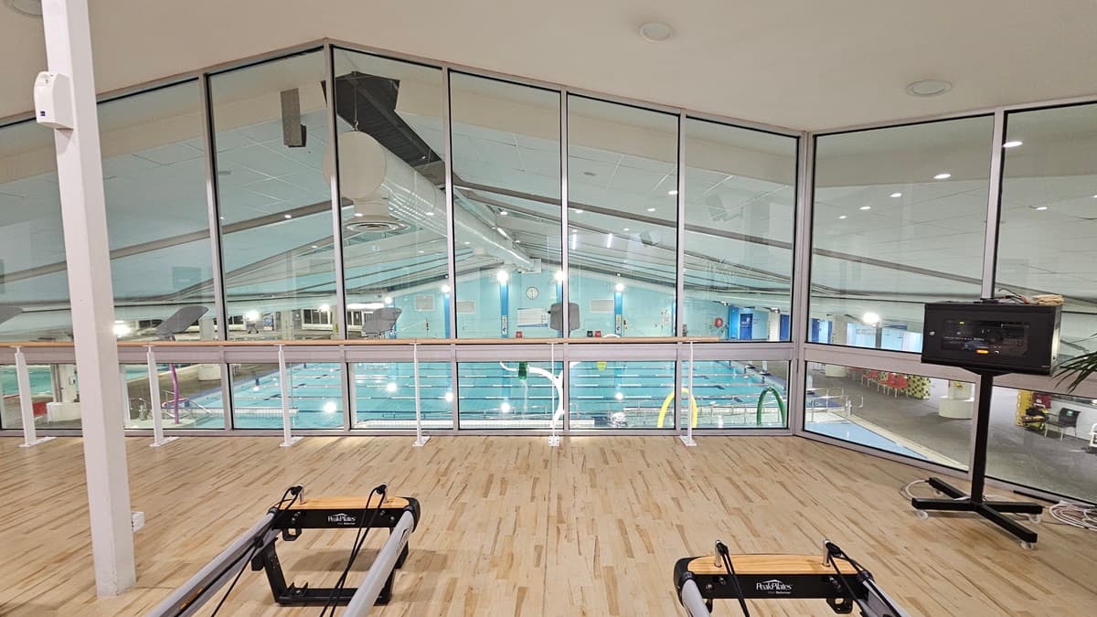 An indoor gym displays Pilates reformers arranged on the left, with a manual glass door entrance adjacent. To the left, there is a rack, a rubbish bin, and large indoor plants placed next to a chair. The white ceiling is fitted with recessed lights, ceiling fans, and air conditioning. The floor is polished timber. Another display rack is located near a black door further inside. The wall on the left features a mirror reflecting the interior.
