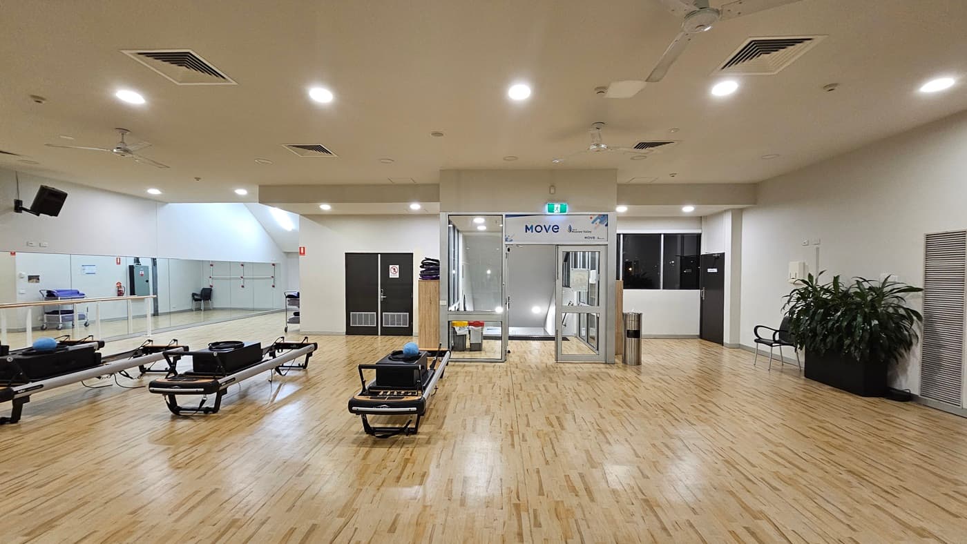 An indoor gym displays Pilates reformers arranged on the left, with a manual glass door entrance adjacent. To the left, there is a rack, a rubbish bin, and large indoor plants placed next to a chair. The white ceiling is fitted with recessed lights, ceiling fans, and air conditioning. The floor is polished timber. Another display rack is located near a black door further inside. The wall on the left features a mirror reflecting the interior.