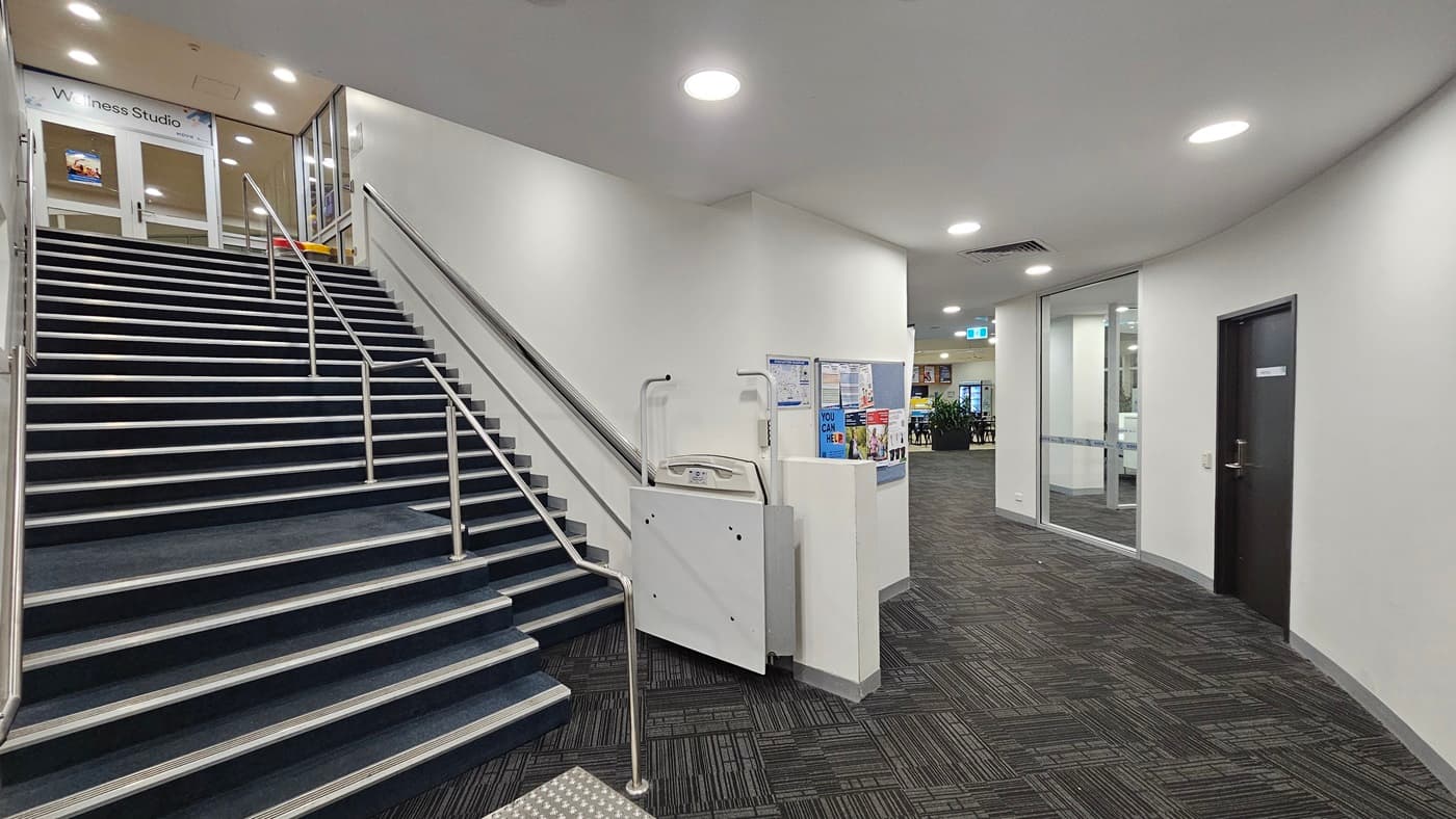 An interior hallway is carpeted with a dark patterned design and features a staircase on the left, which includes metal railings and contrasting edge strips along with truncated domes. Adjacent to the staircase is a white information stand. The walls are white and equipped with notice boards. The white ceiling incorporates air conditioning and recessed lighting. To the right, a glass door and a wooden door are positioned next to each other.