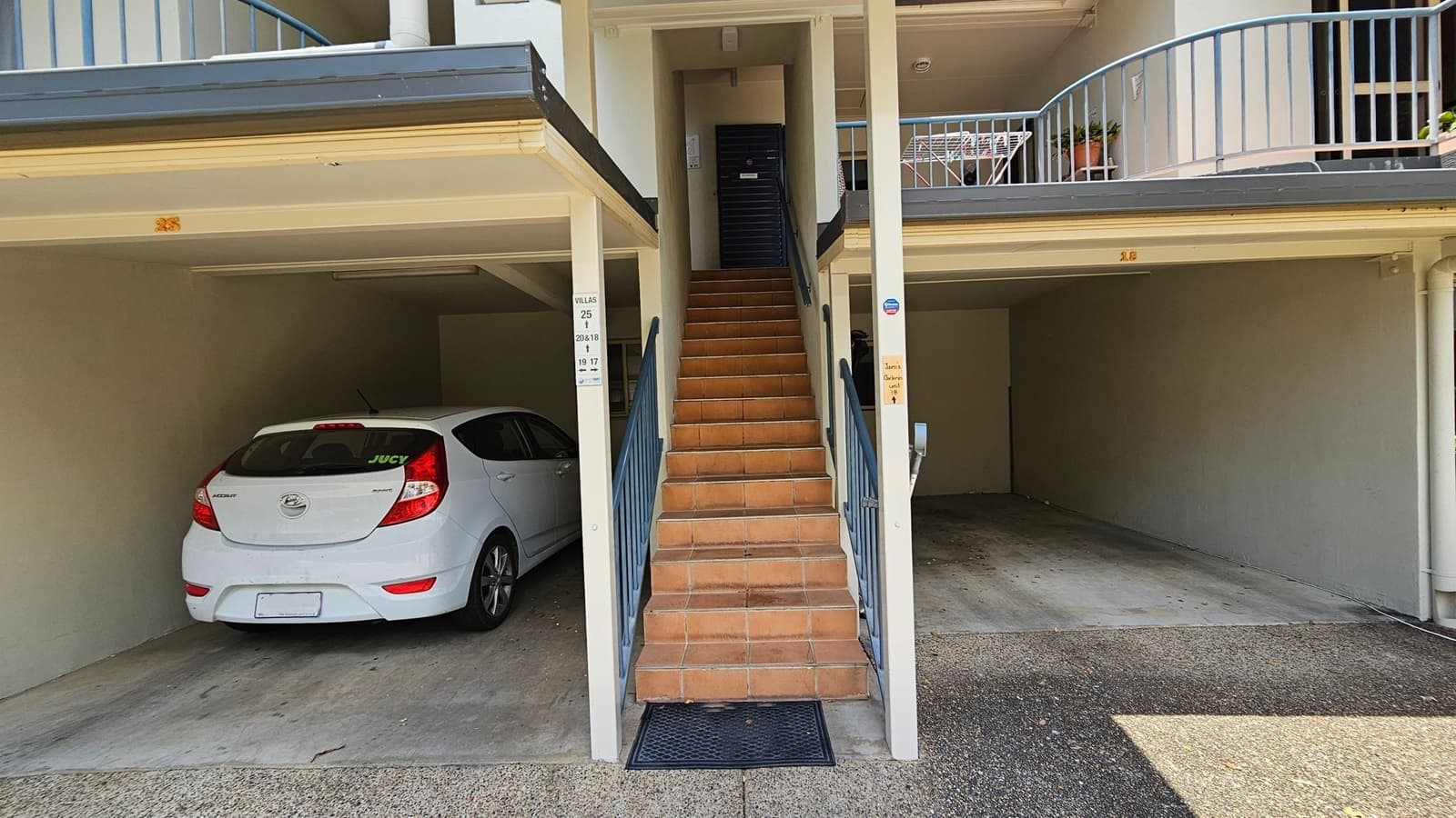 A white Hyundai car is parked in a covered space on the left. In the centre, a set of terracotta-coloured stairs with blue railings leads up to a black door. On the right, there is an empty parking space. Above, a balcony with a blue railing holds a small table and potted plants. The ground is a mix of concrete and gravel.