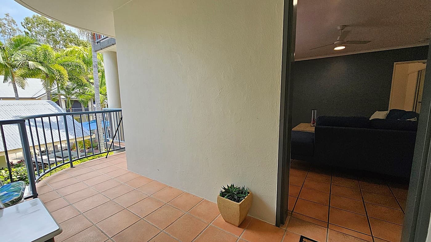 A balcony with terracotta tiles and a metal railing on the left overlooks a garden with palm trees and a building with a corrugated metal roof. A small potted plant sits on the floor near the wall. On the right, an open door leads into a dimly lit room with a dark sofa, a side table, and a ceiling fan.