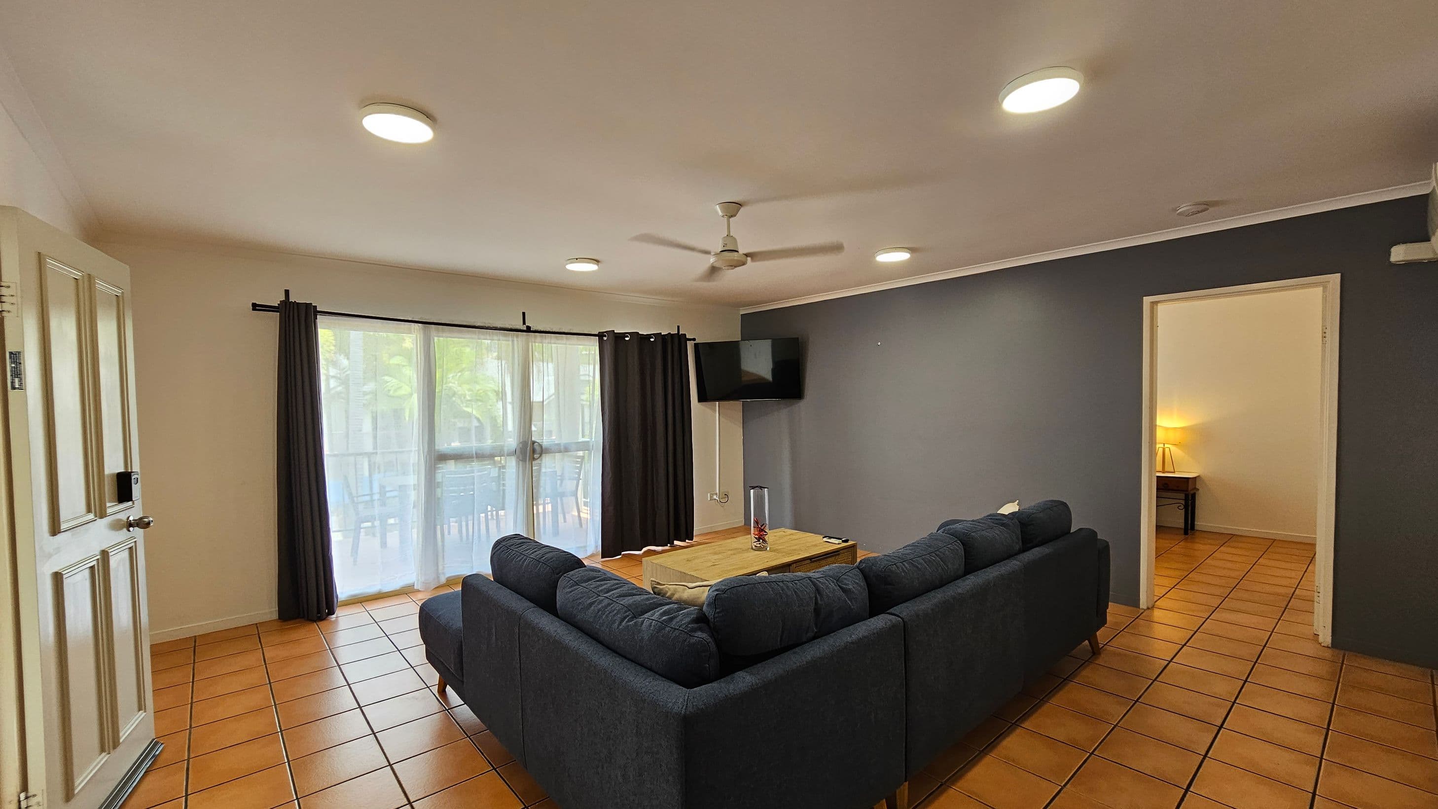A living room with terracotta tiled flooring and a white ceiling with recessed lights. On the left, there's a partially open door and large windows with sheer and dark curtains. In the centre, a dark grey L-shaped sofa surrounds a wooden coffee table with a decorative vase. A wall-mounted TV is on the right wall, which is painted grey. An open doorway leads to another room with a lit lamp visible. A ceiling fan is centred above the seating area.